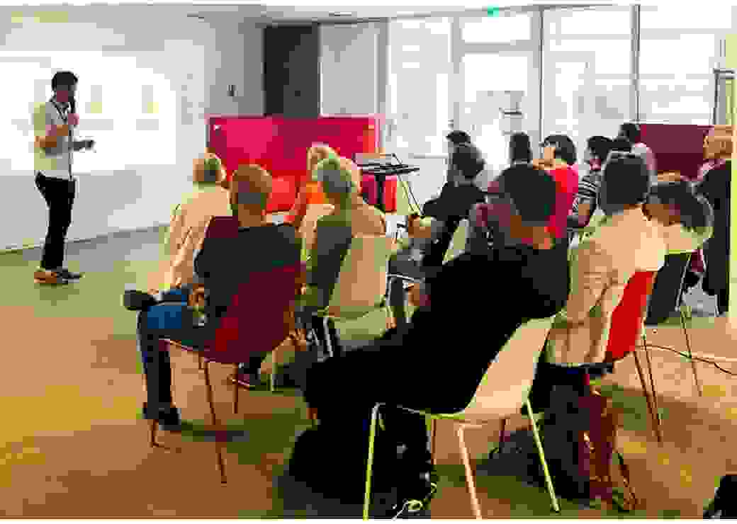 Dans une salle, des personnes assises écoute un homme qui parle au micro, devant une projection de graphiques