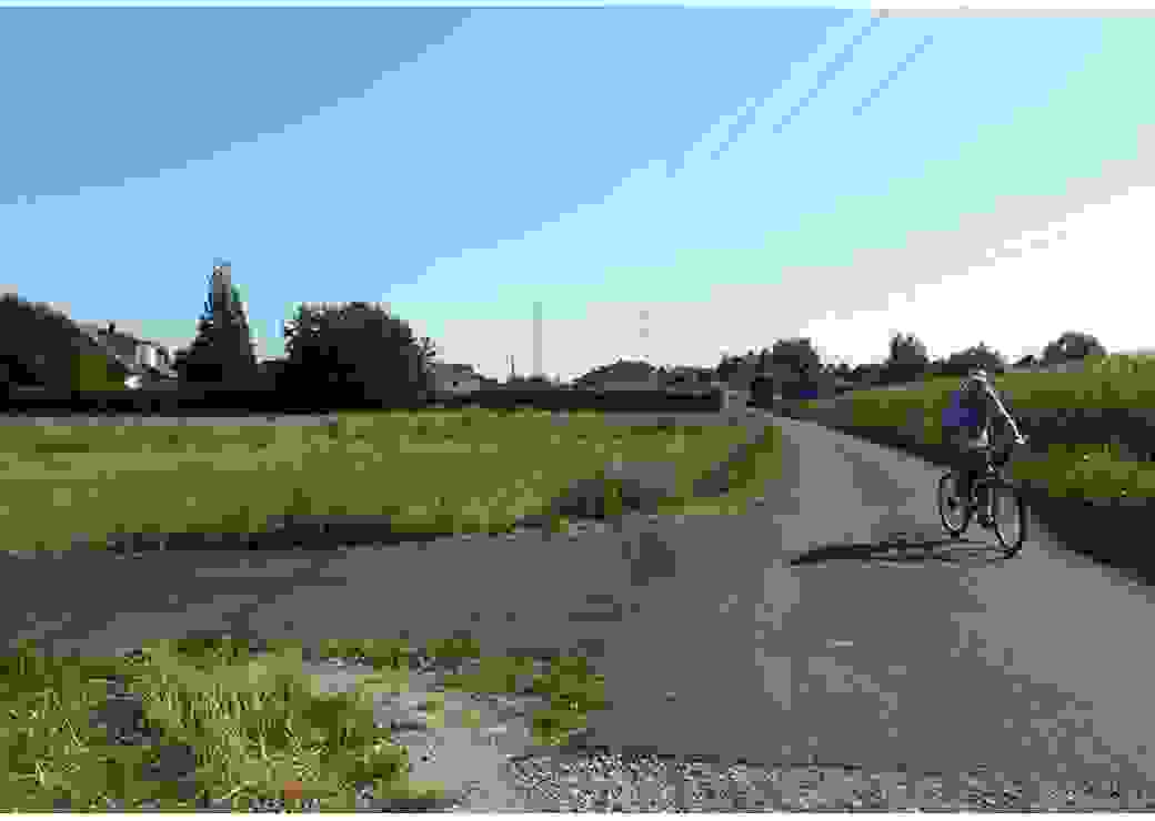 Un homme circule à vélo sur un chemin bordé de champs et de prairie, il avance vers le photographe. Au loin, on distingue une zone pavillonnaire