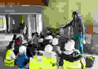 Un groupe d'enfants coiffés de casques de chantier est assis devant un homme qui tient des rameaux dans sa main. Ils sont dans un bâtiment en chantier