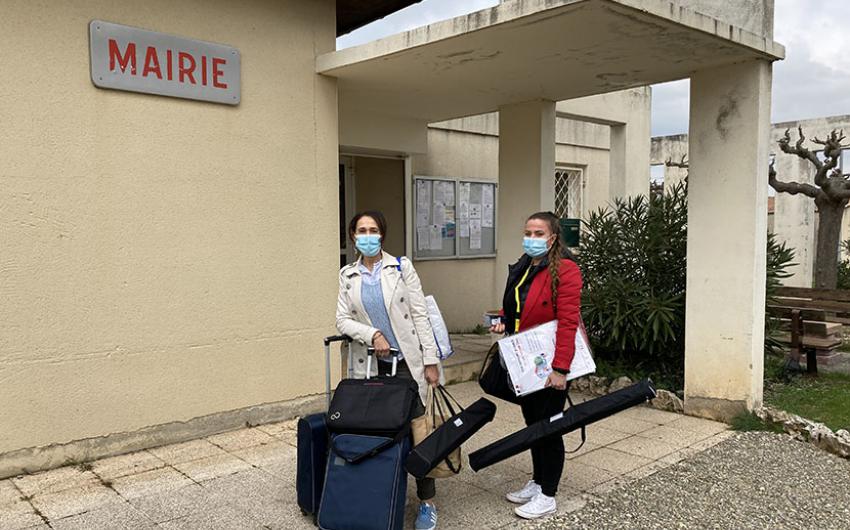 Devant un bâtiment affichant un panneau "mairie", deux femmes chargées de bagages posent pour le photographe