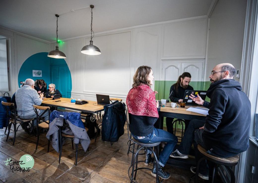 Six personnes sont assises devant des tables, certaines discutent, d'autres travaillent sur des ordinateurs