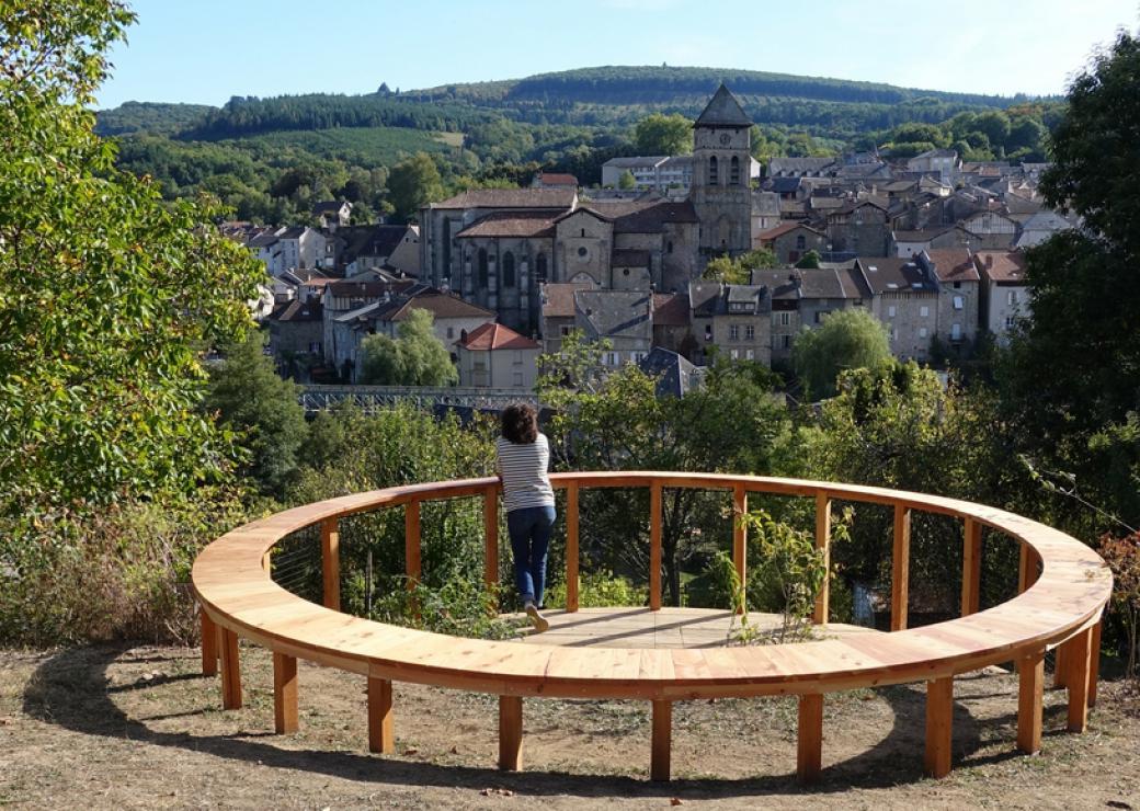 Sur une colline, une personne est appuyée à une construction en bois circulaire et regarde le village situé sur la colline opposée