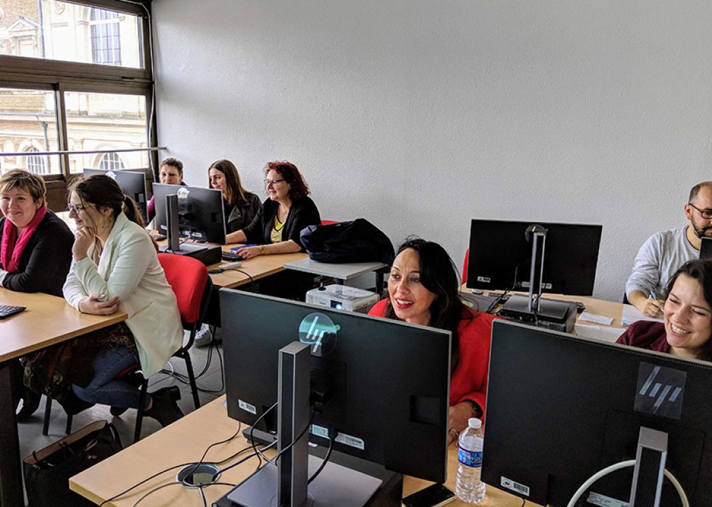 Dans une salle de classe, 8 adultes, principalement des femmes, sont attablés devant des écrans d'ordinateurs