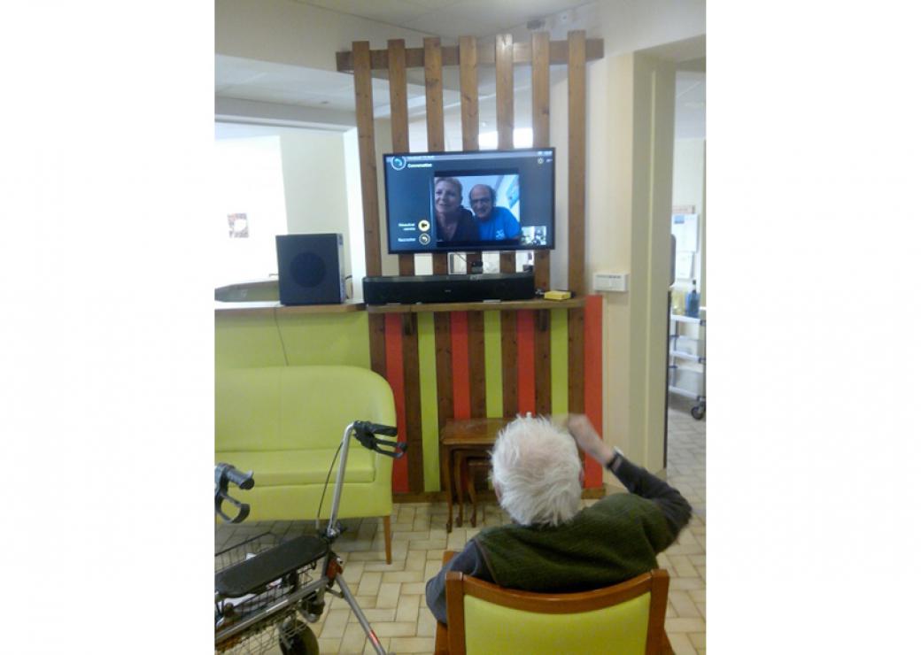 Une personne âgée est installée devant un téléviseur dont l'écran montre deux personnes qui semblent être en visioconférence