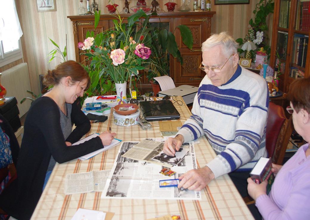 Dans le salon d'une maison, un homme et une femme âgés exposent sur la table des photos et des coupures de presse. Face à eux, une jeune femme prend des notes.