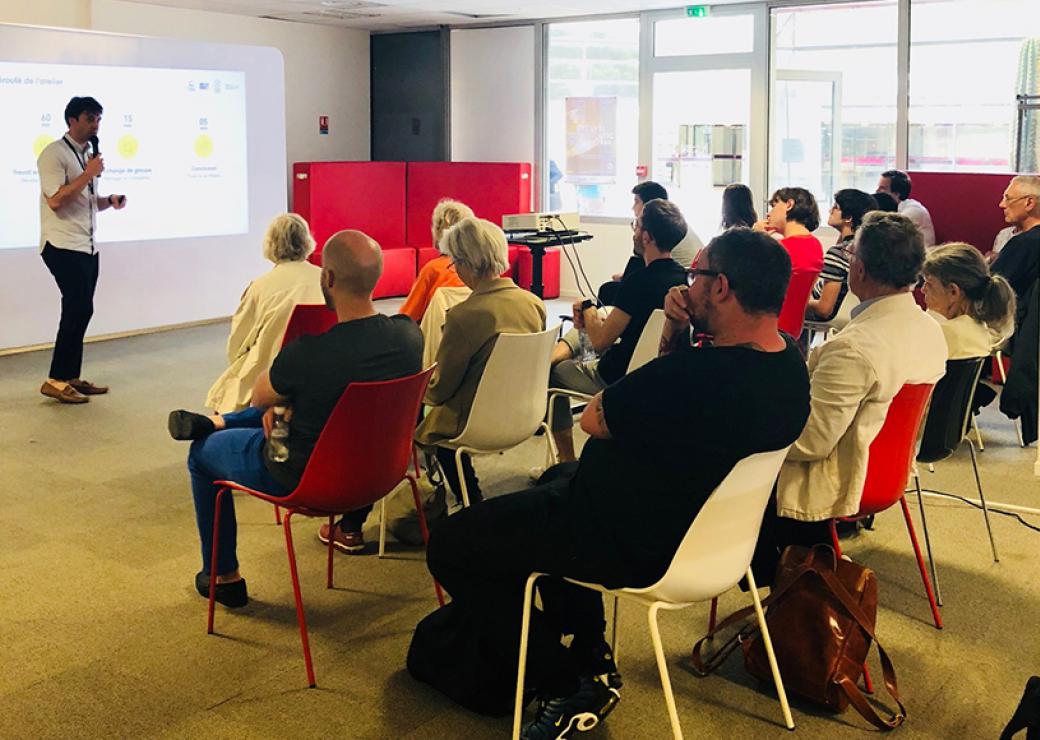 Dans une salle, des personnes assises écoute un homme qui parle au micro, devant une projection de graphiques