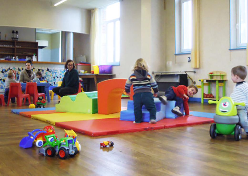 Dans une salle éclairée par trois fenêtres, des enfants en bas âge jouent sur des tapis colorés, sous la surveillance de 2 adultes