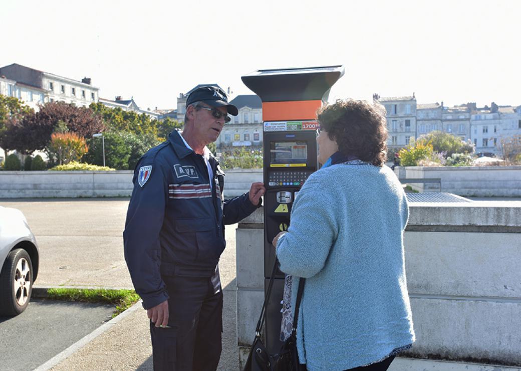 Un policier municipal en uniforme et une dame parlent devant une borne de parcmètre