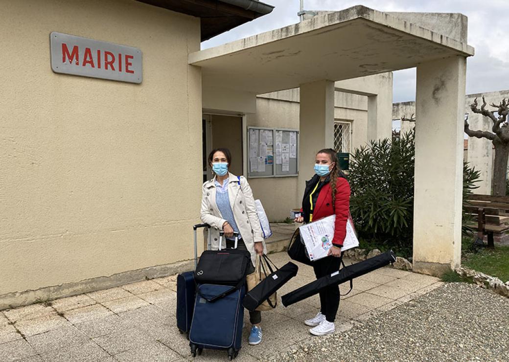 Devant un bâtiment affichant un panneau "mairie", deux femmes chargées de bagages posent pour le photographe