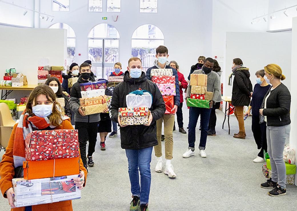 dans une grande salle, des personnes avancent face caméra, les bras chargés de paquets cadeaux