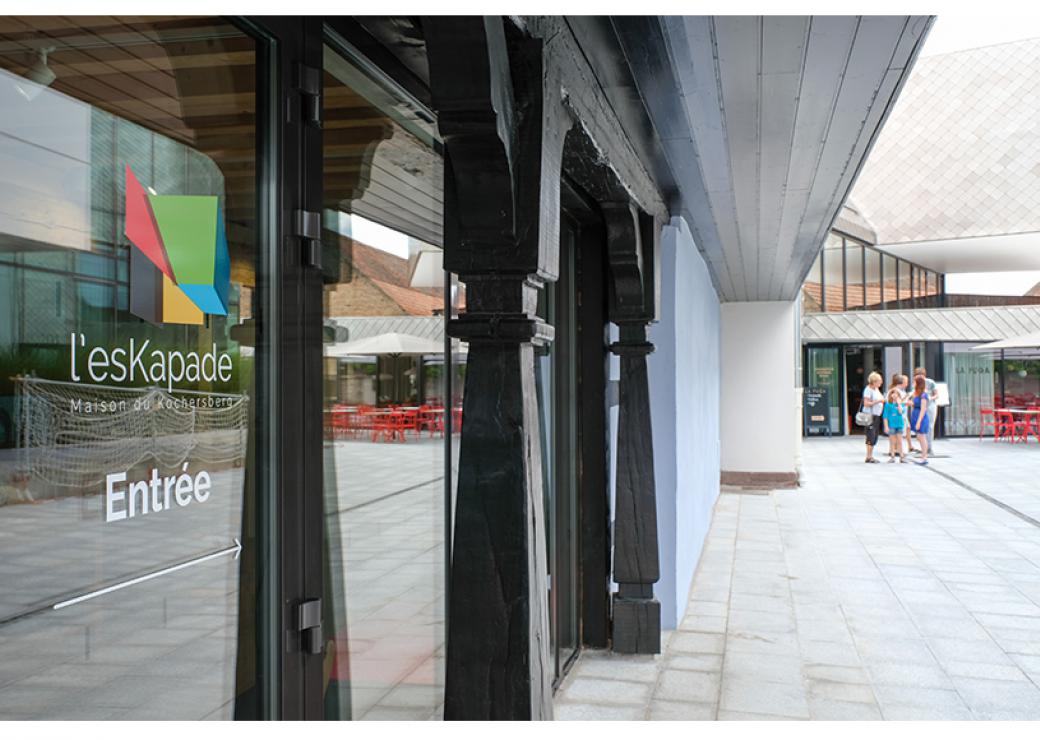 plan latéral d'une vitrine sur laquelle on peut lire "l'Eskapade", maison du Kochersberg. En arrière plan, un groupe de 4 ou 5 personnes