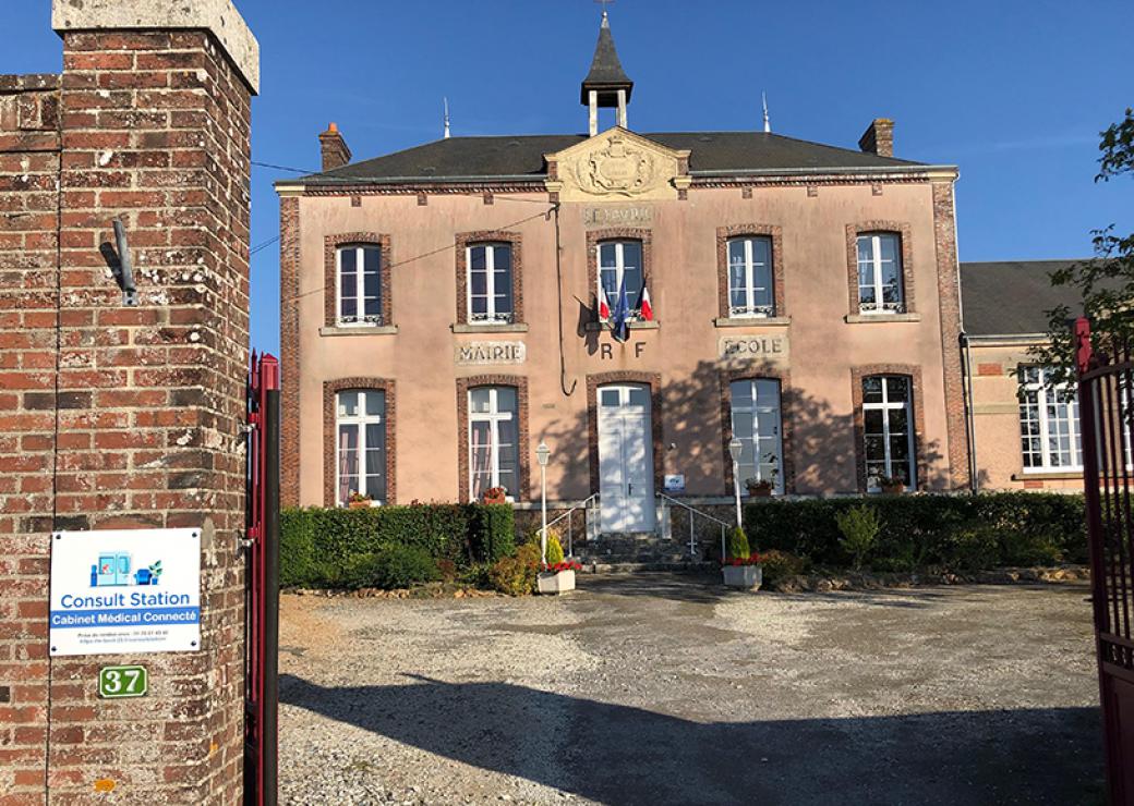 Photo plan large d'un bâtiment de deux étages, en façade duquel on peut lire "mairie, RF, école". Sur l'une des piles du portail que l'on devine dans l'encadrement de la photo, on peut "consult station, cabinet médical connecté"