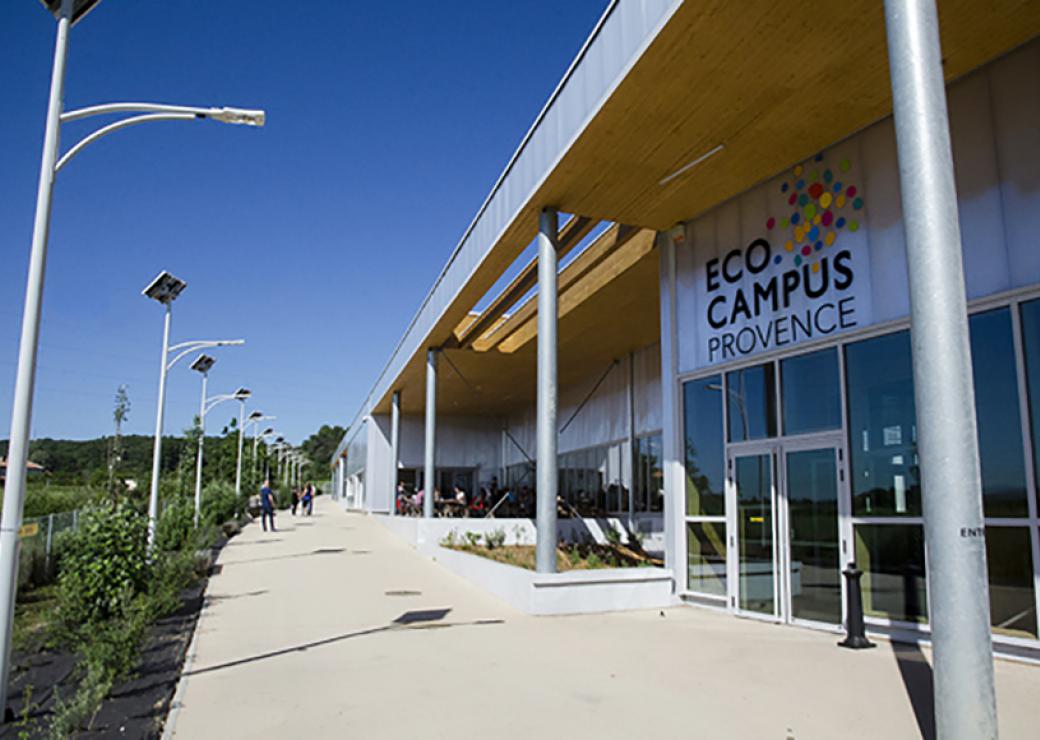 Une rangée de réverbères à gauche délimitent le chemin qui borde une façade moderne sur laquelle on peut lire "Eco campus Provence"