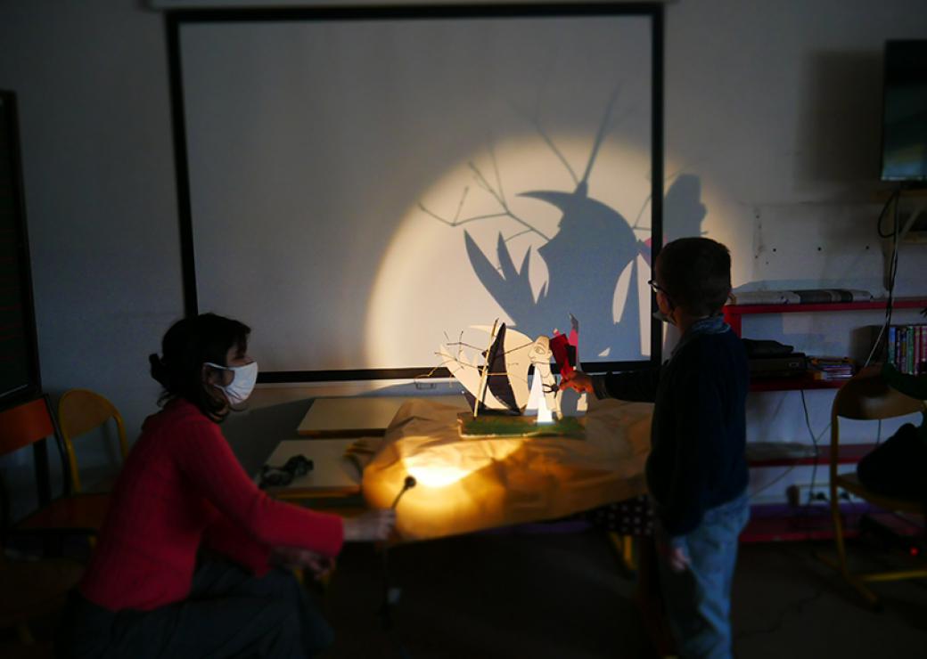 Dans une pièce peu éclairée, une femme dirige le faisceau d'une lampe vers une construction que tient un enfant. Sur le mur du fond, se projette l'ombre de la construction.