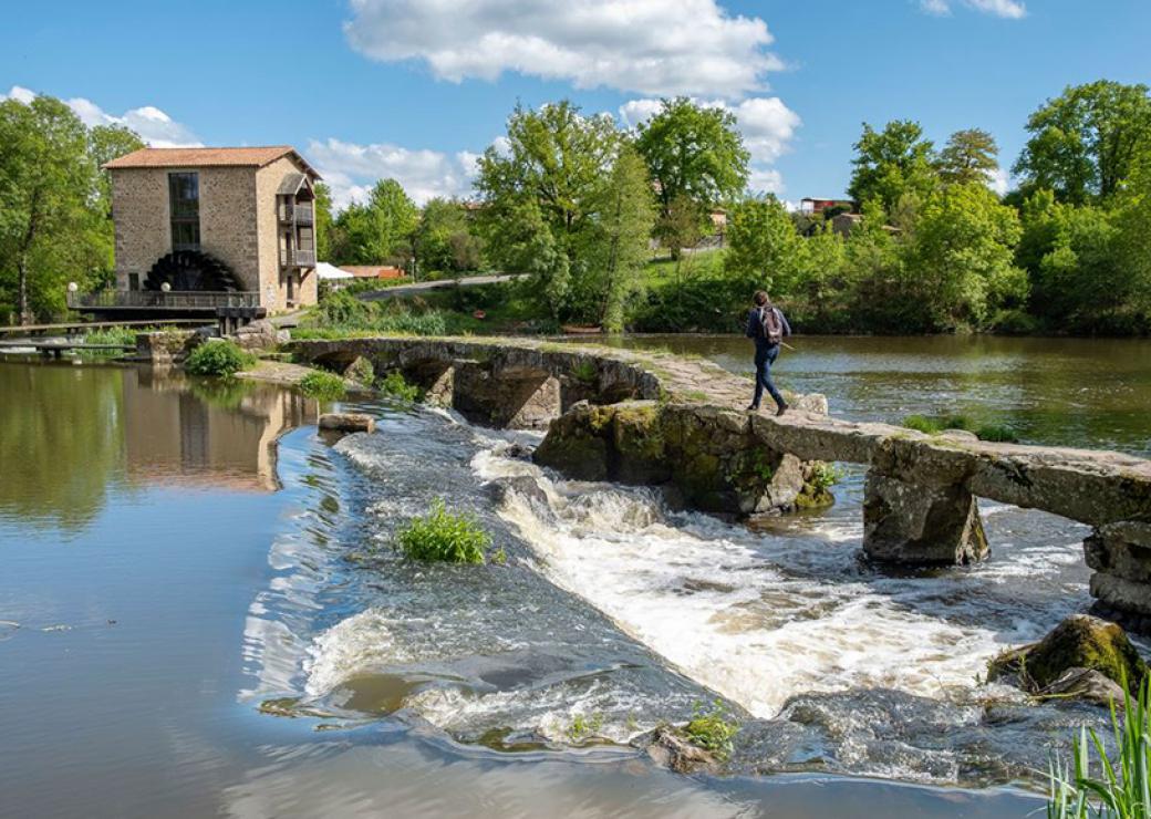 Une personne chemine sur une passerelle de pierre au dessus d'une rivière, en direction d'un moulin sur la berge opposée