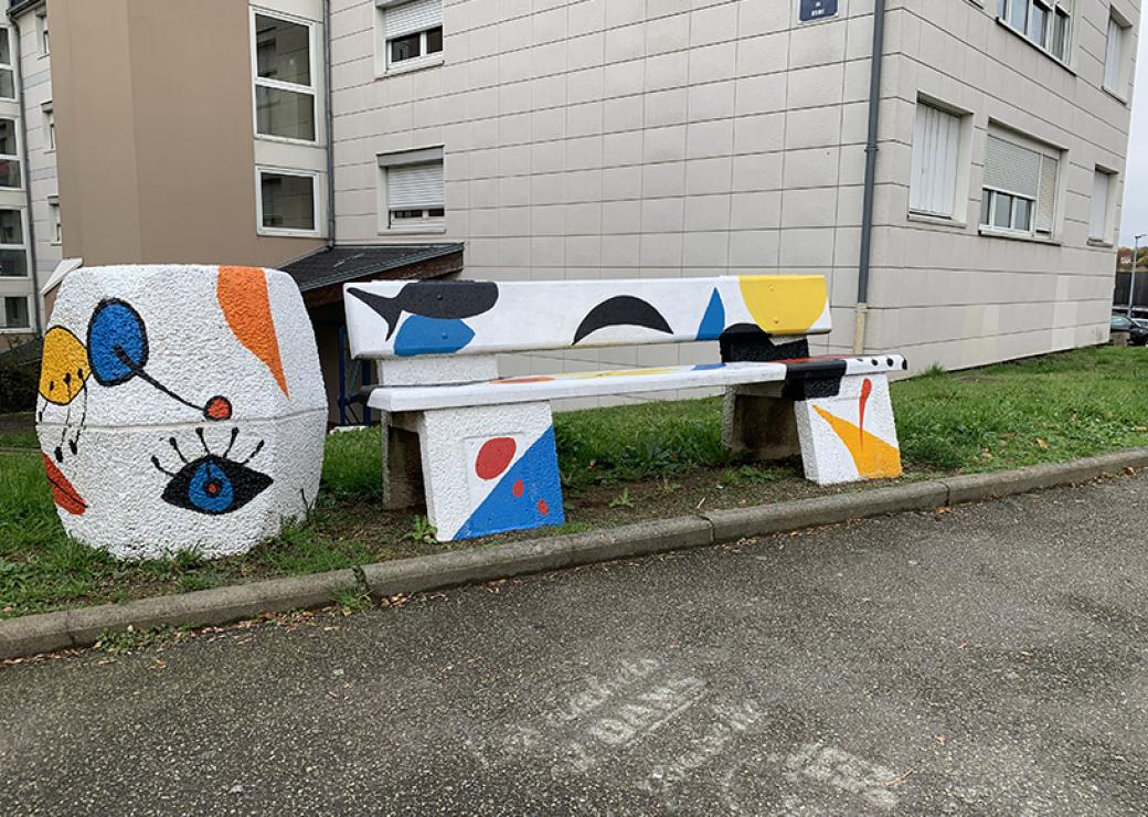 En pied d'immeuble, un banc et un pot de fleur sont décorés avec des motifs colorés, dans le style du peinte miro