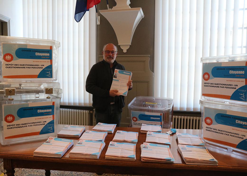 Un homme se tient devant un bureau couverts d'enveloppes et de cartons sur lesquels on peut lire "grande consultation citoyenne"