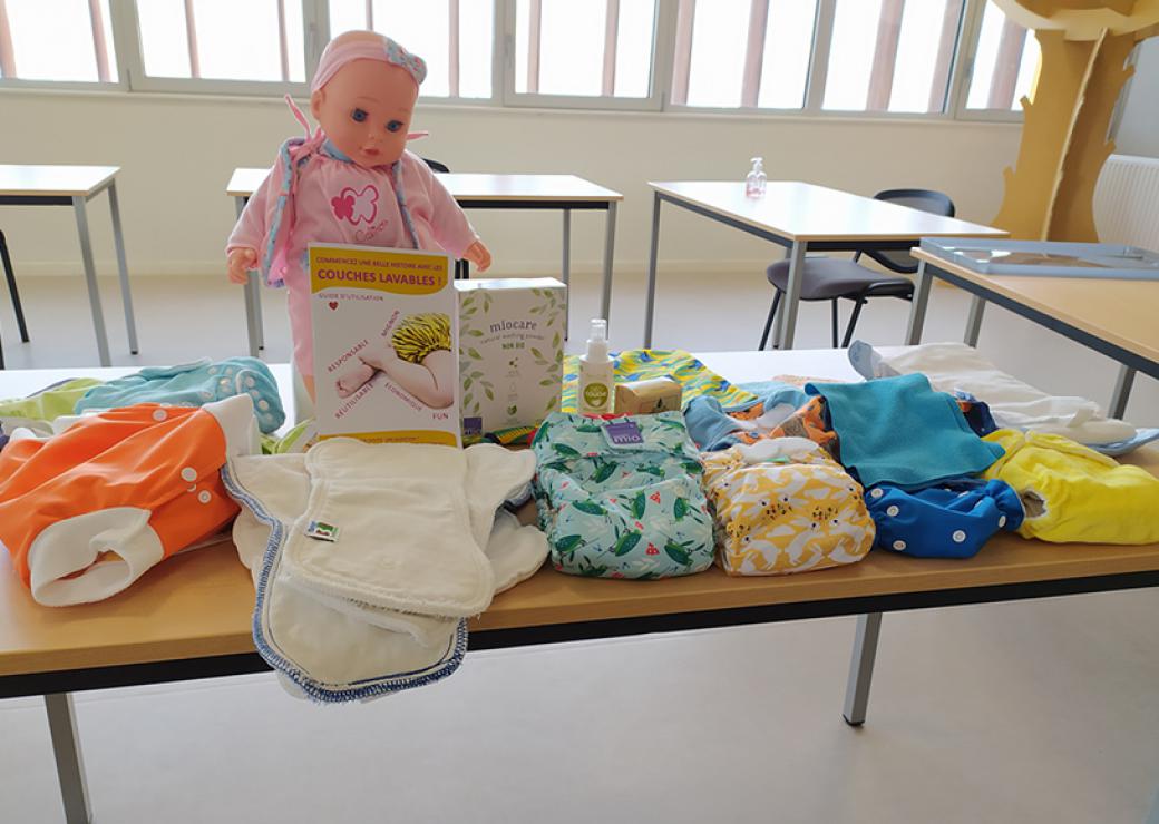 Sur une table, différents modèles de couches sont exposés, et un poupon de plastique surmonte le tout