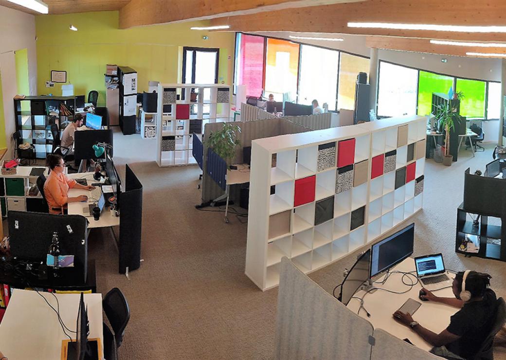Plan large d'une vaste salle aux grandes vitres colorées. C'est un open space où l'on compte 4 personnes et une dizaine d'espace de travail