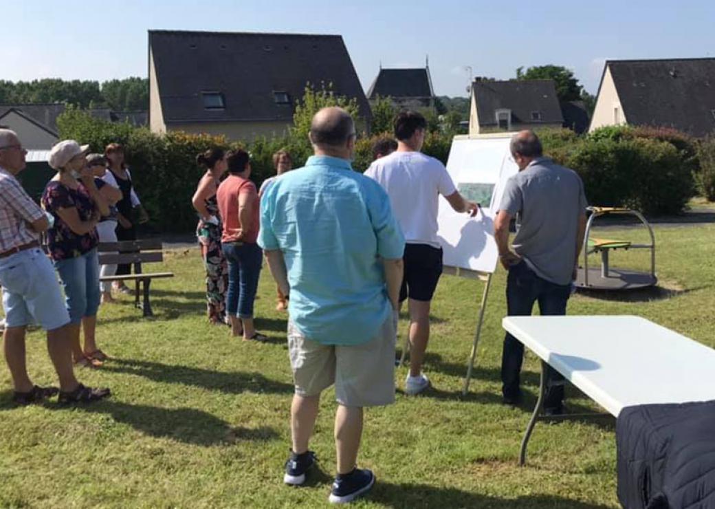 Des personnes sont réunies sur une pelouse, dans un lotissement, autour d'un homme qui dessine sur un tableau de feuilles blanches