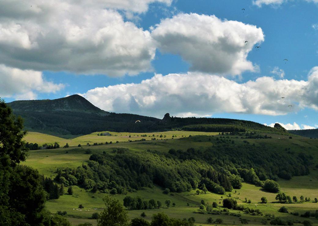 Vue d'un paysage vallonné, alternant clairière et bois. Dans le ciel, on distingue 6 voiles de parapente