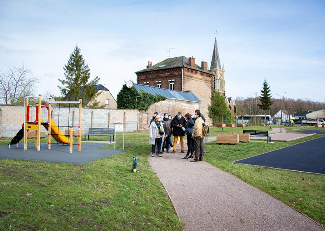 Dans un jardin public, à droite des jeux pour enfants, au milieu un sentier goudronné sur lequel se situe un groupe de personnes portant des masques chirurgicaux