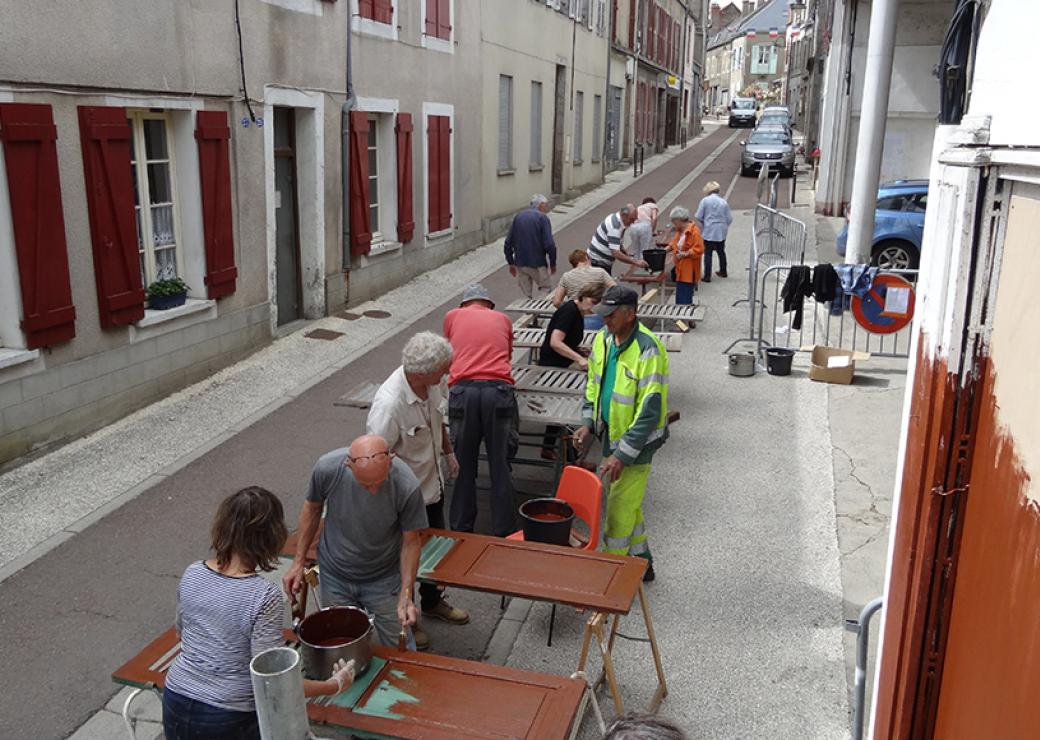 Dans une rue de village, une douzaine de personnes est affairée à peindre des volets de couleur marron