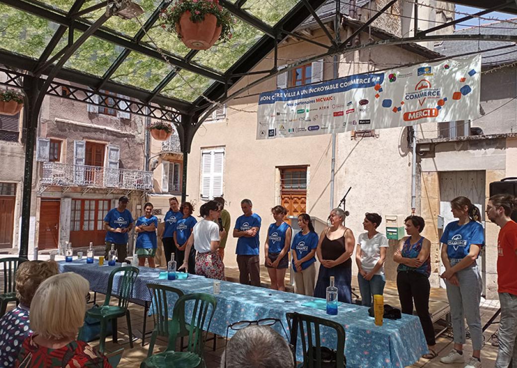 Sur une ^lace de village, des personnes se tiennent en ligne devant une grande table. Ils semblent faire face à un public.