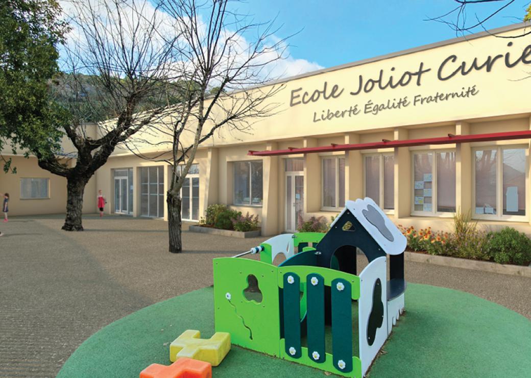 Vue d'une cours d'école: une aire de jeux colorée, et un fronton sur lequel on peut lire Ecole Joliot Curie Liberté égalité fraternité
