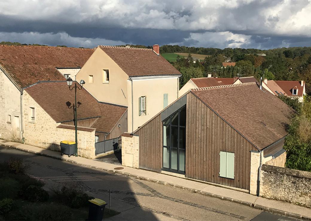 Dans un village, des maisons traditionnelles et un bâtiment rénové avec une grande fenêtre et un bardage de bois sur la façade