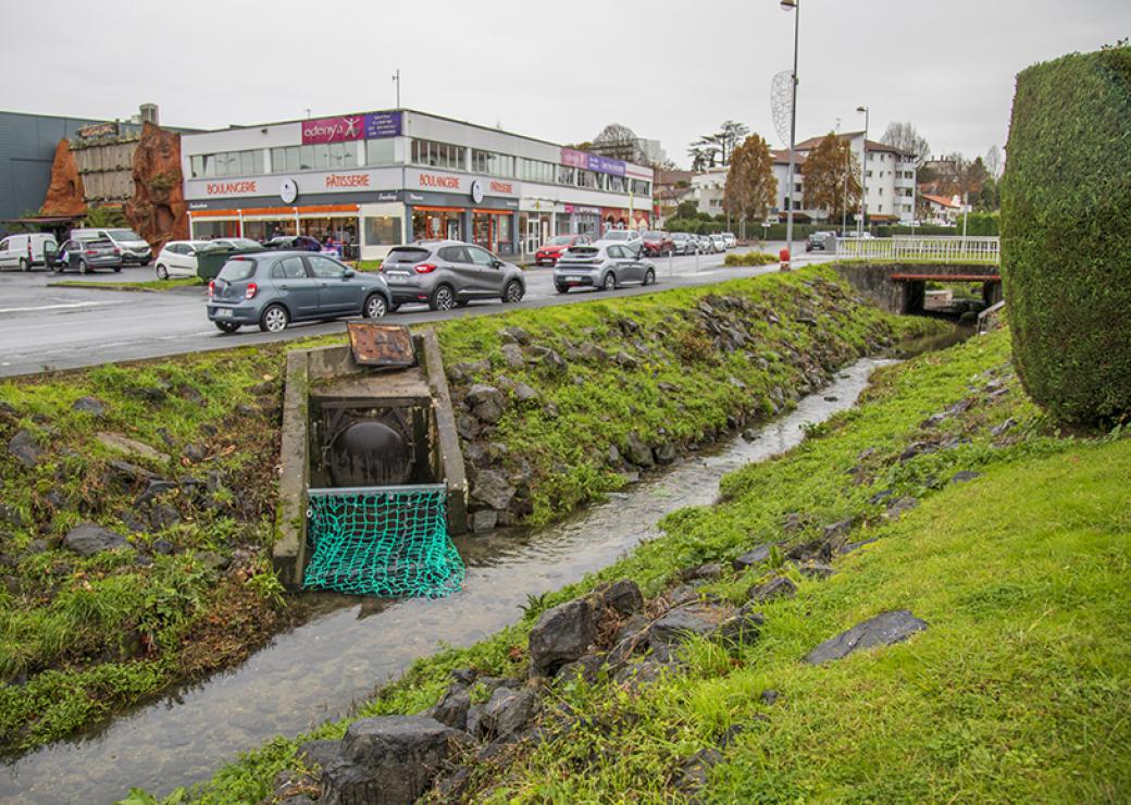 En contrebas d'une route, un ruisseau danss lequel vient se jeter une grosse canalisation équipée d'un filet vert