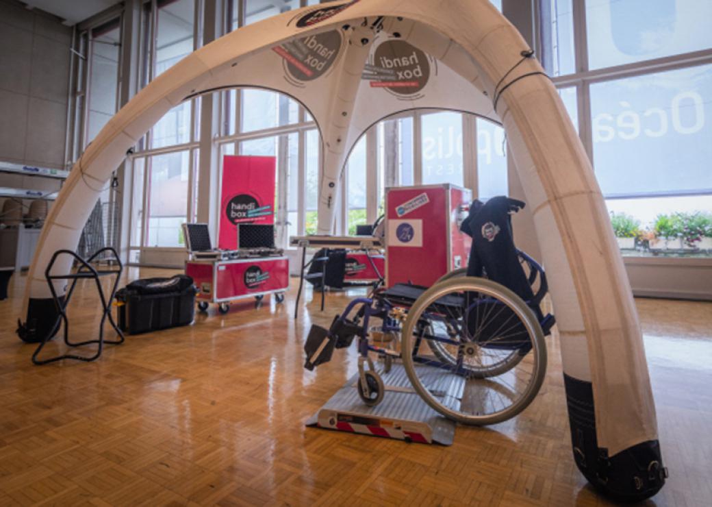 Dans une très grande salle, sous un chapiteau ouverts sur les côtés, sont présentés un fauteuil roulant et des grandes caisses rouges