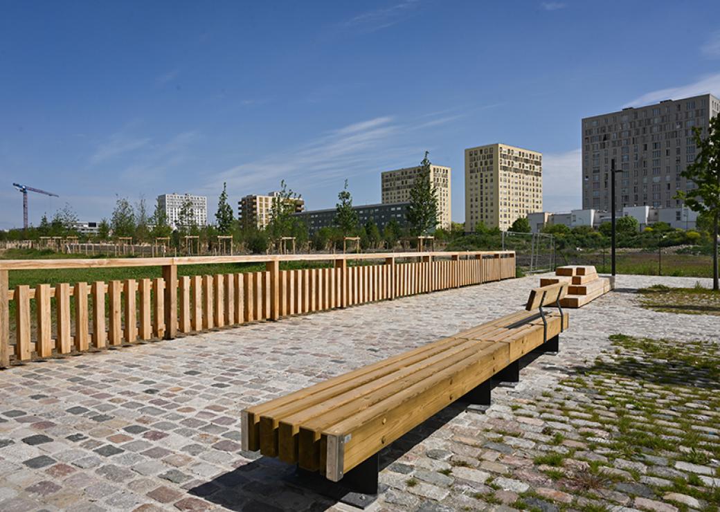 Vue sur un espace public pavé et meublé de bancs de bois. En arrière-plan, 6 grands immeubles