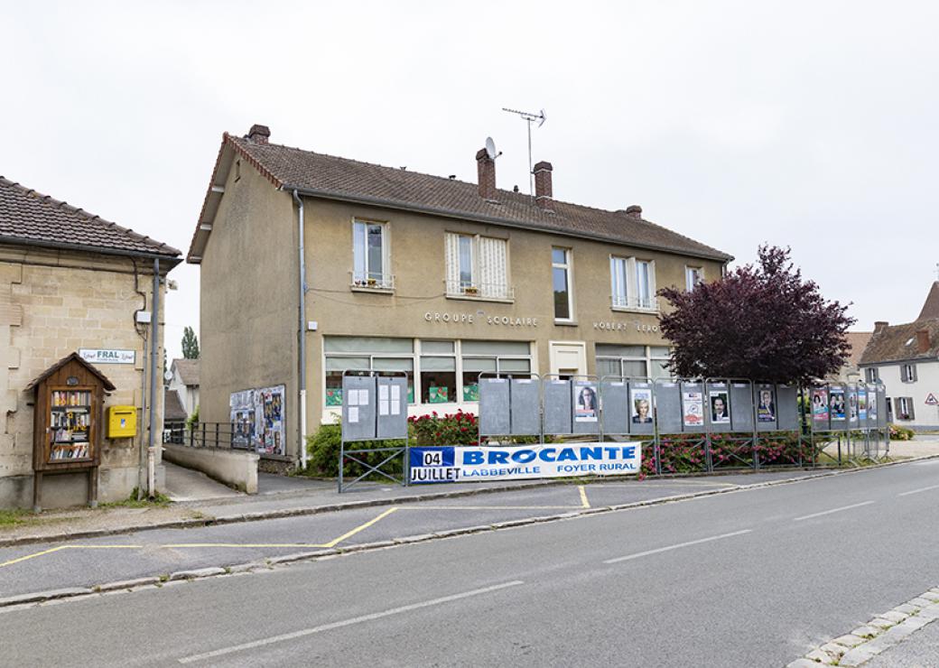 Depuis le bord d'une route, photo d'un bâtiment sur lequel on peut lire groupe scolaire. Une banderole indique aussi Brocante Labbeville Foyer rural 04 juillet