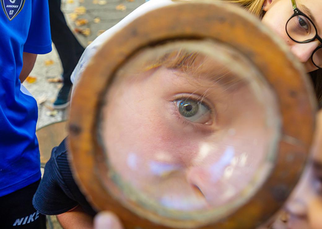 Un oeil d'enfant en très gros plan, vu au travers d'une loupe ou en reflet dans un miroir. Un autre enfant, au second plan, semble se pencher vers le premier enfant.
