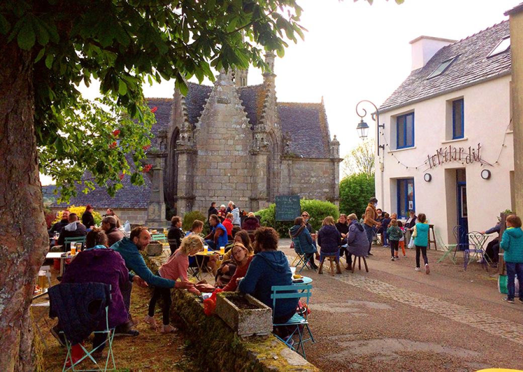 Des gens sont attablés à des tables sur une terrase de café. Un marronnier borde la terrasse, au fond se détache un édifice religieux