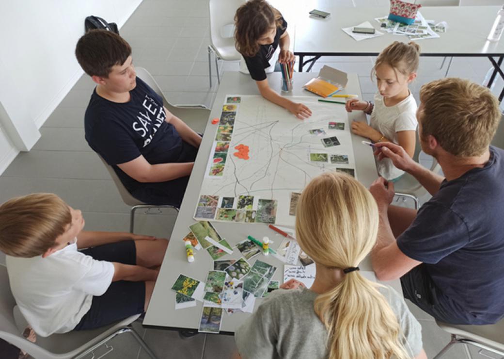 Vue du dessus, une table sur laquelle sont posés une carte et des photos. 5 personnes, enfants et adultes, se tiennent autour de la table