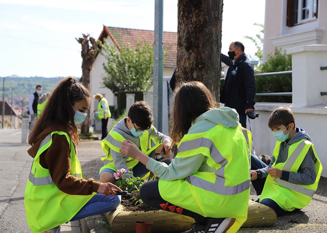 Des enfants habillés de gilets jaunes plantent des végétaux au pied d'un arbre sur un trottoir