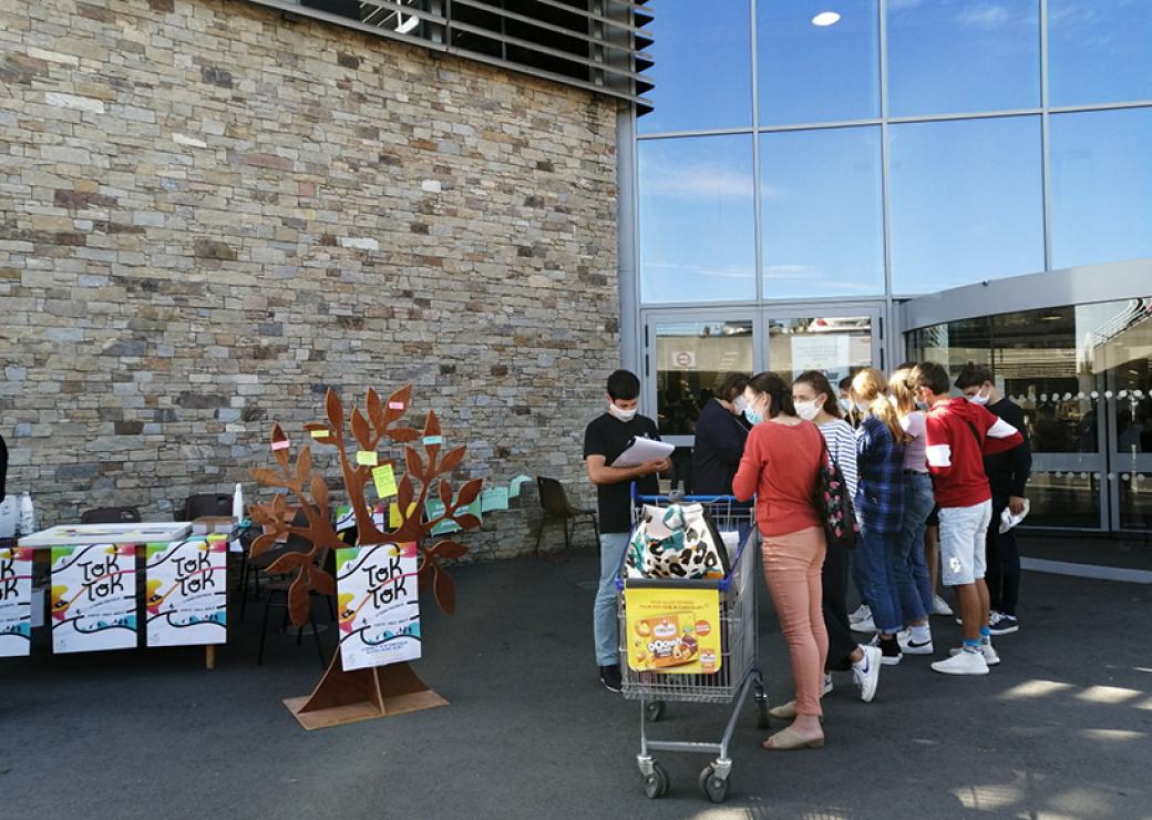 Devant l"entrée d'un supermarché, des jeunes sont regroupés et semblent discuter. Des affiches sont apposée sur une table. On peut lire TokTok sur ces affiches