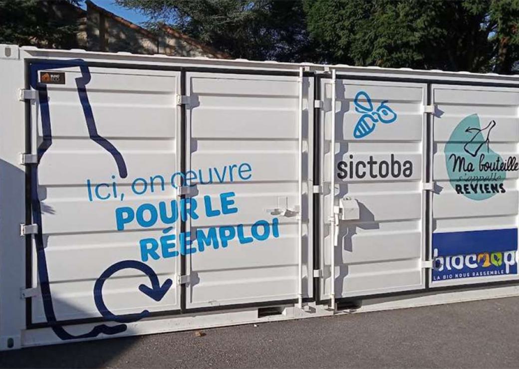 Un container blanc sur lequel on peut lire "ici on œuvre pour le réemploi", "sictoba" et "ma bouteille s'appelle reviens"