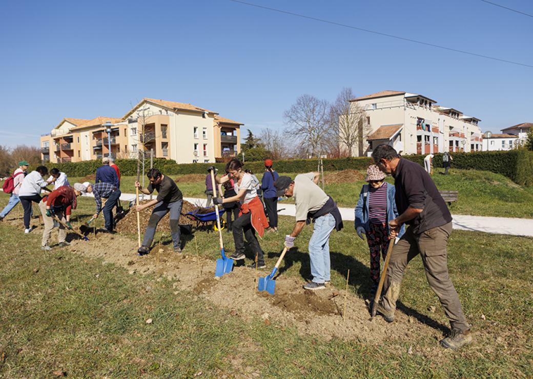 En ligne, une vingtaine de personnes creusent la terre à l'aide d'une pelle. Au fond, des petits immeubles d'habitation