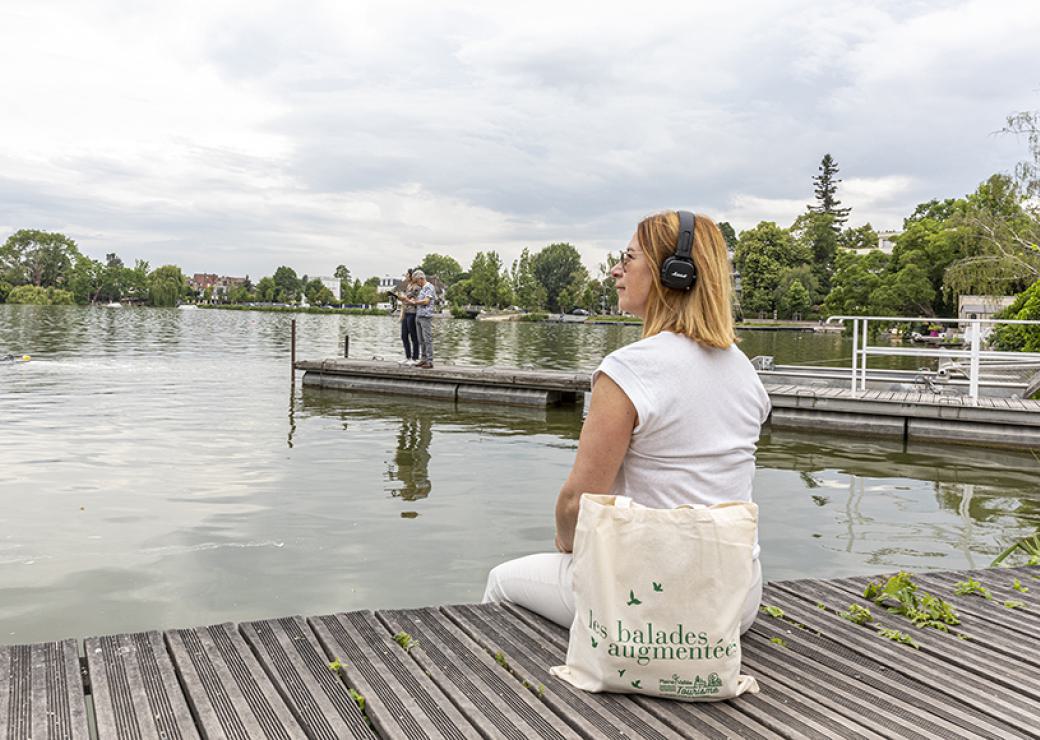 Une femme vetue de blanc est assise sur un ponton de bois,, face à une étendue d'eau. Elle porte un casque audio sur les oreilles