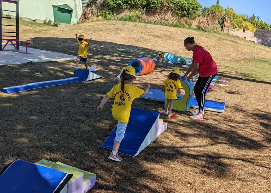 En plein air, trois enfants évoluent sur un parcours d'activité, sous la surveillance d'une jeune femme