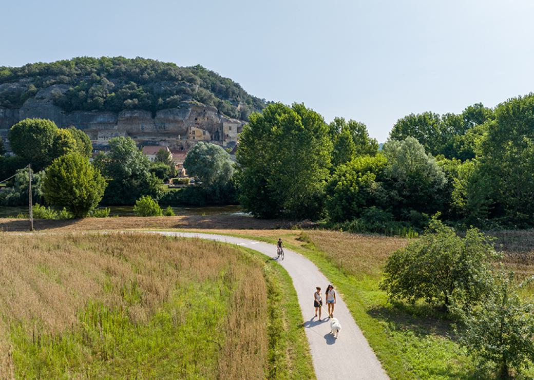 Vu d'en haut, un chemin sur lequel se promènent deux personnes et leur chien et une personne à vélo. Paysage de campagne