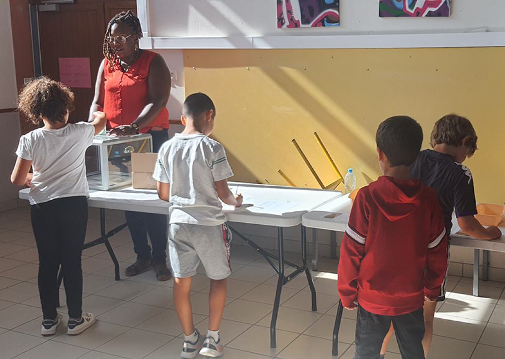 Une jeune femme tient une urne dans laquelle un enfant dépose un bulletin. Trois autres enfants sont en train d'émarger