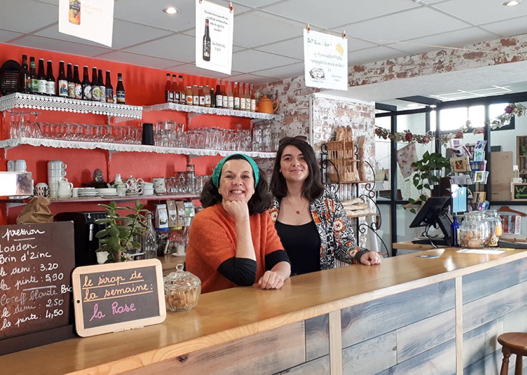 Deux femmes se tiennent derrière le comptoir d'un bar et regardent en souriant la personne qui prend la photo