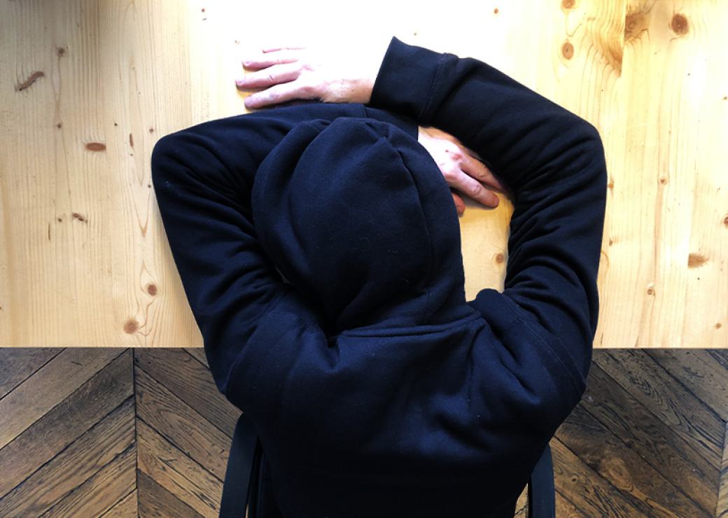 Vue de haut d'une personne, capuche sur la tête et tête reposant sur la table, avec les bras autour