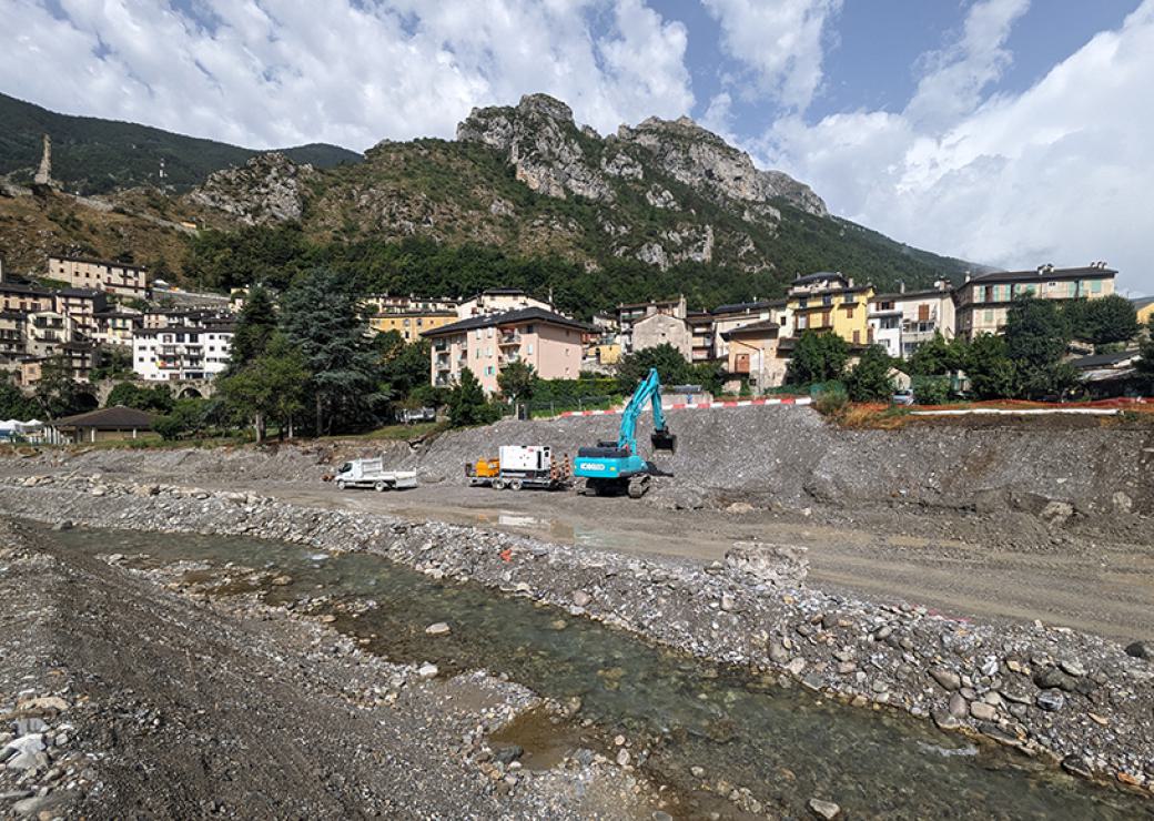 Des engins de travaux sur les berges d'une rivière. En fond, un village bordé par une montagne