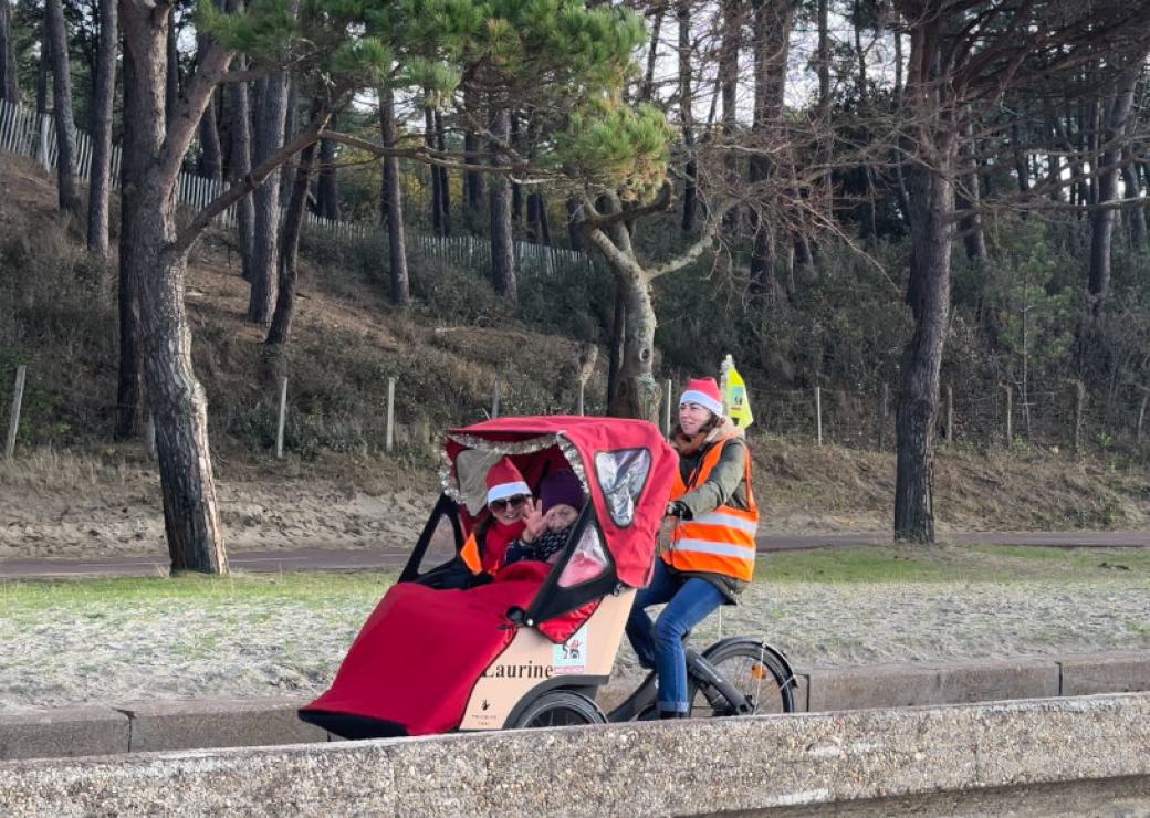 Deux personnes installées dans la cabine d'un vélo triporteur, et une personne qui pédale sur le vélo, sur une route en contrebas d'une forêt de pins