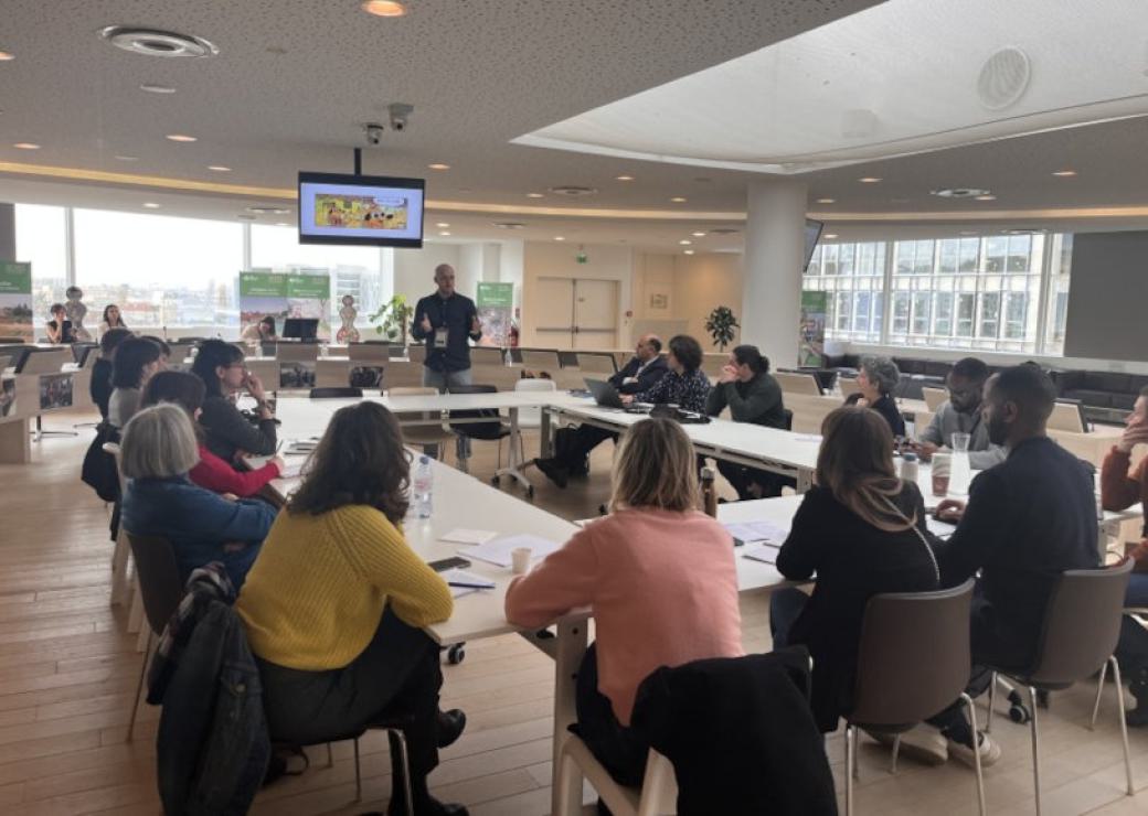 Des personnes assises à des tables disposées en carré, un homme debout devant ces personnes